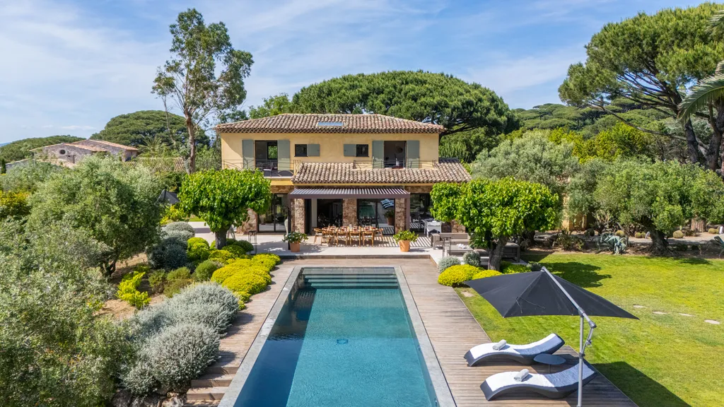 The villa's ochre façade and terrace behind the pool, framed by its Mediterranean garden on a bright day