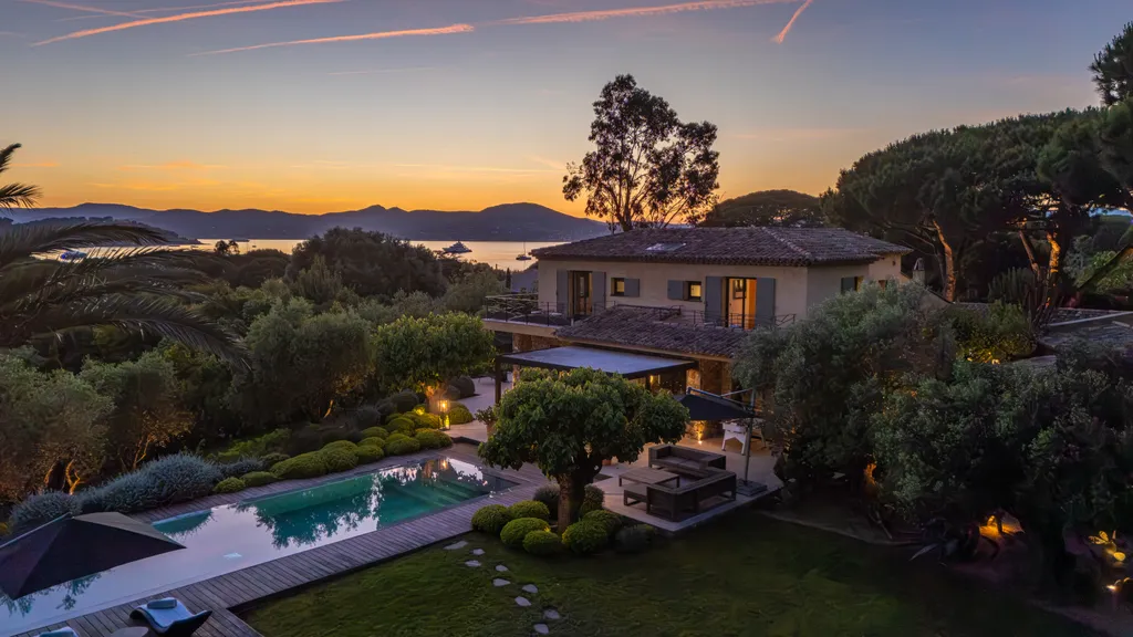 Aerial view at dusk — the lit villa above the bay, the pool glowing in the foreground