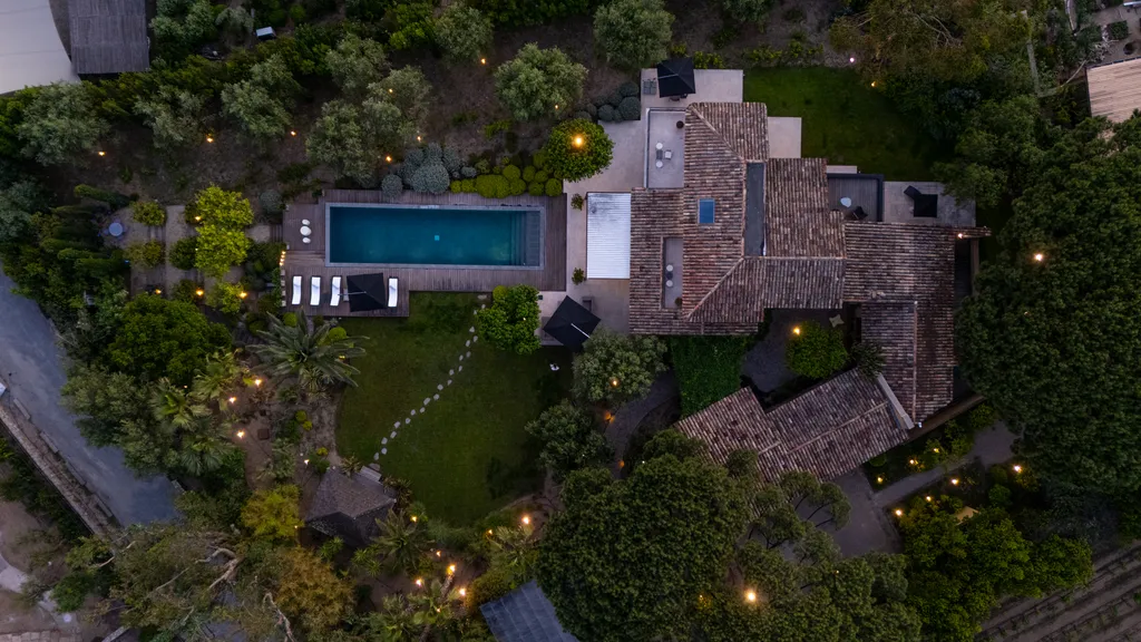 Overhead aerial at dusk — the pool, lawn and tiled roof laced with path lights