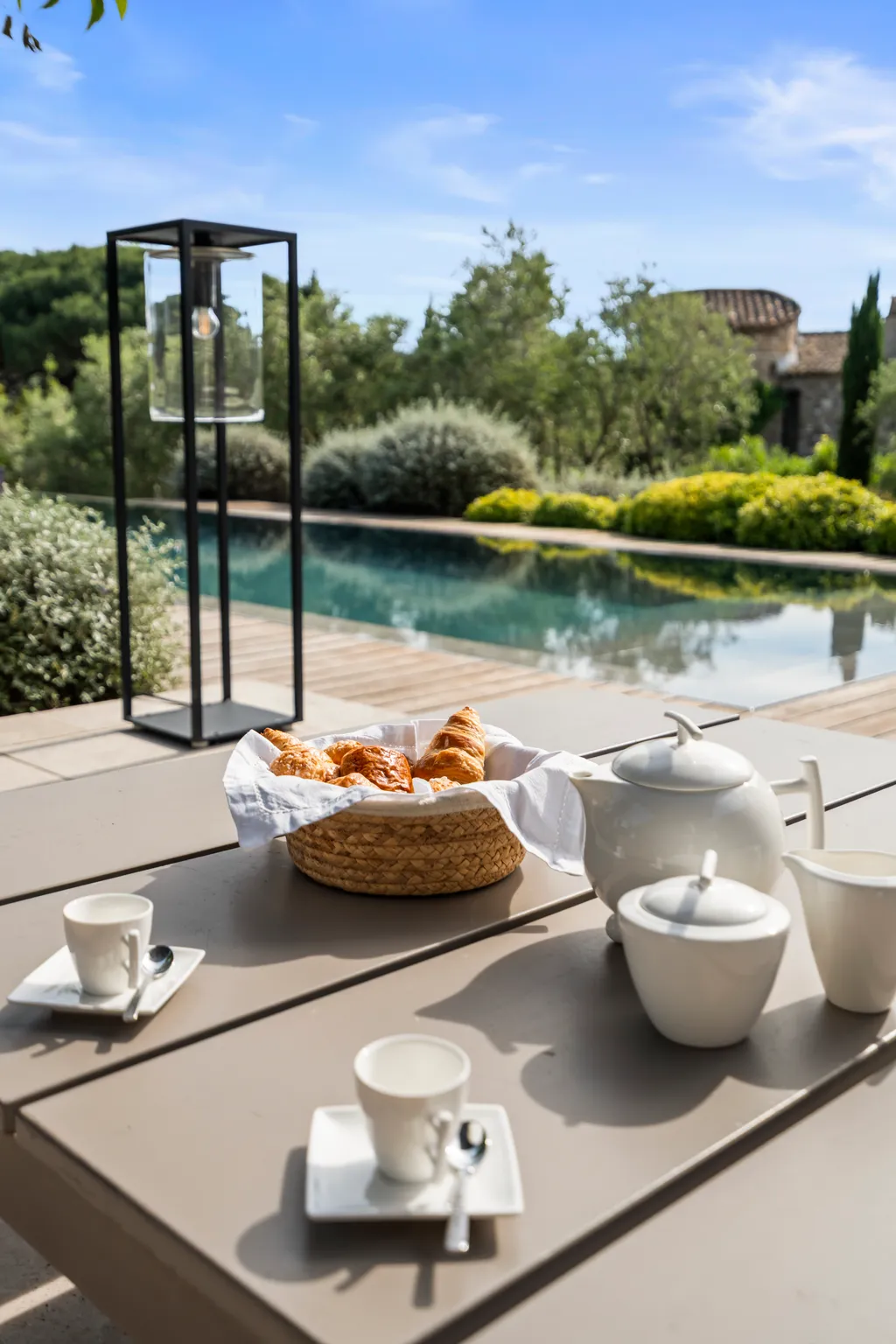 Croissants and coffee on a poolside table, a lantern and the garden behind